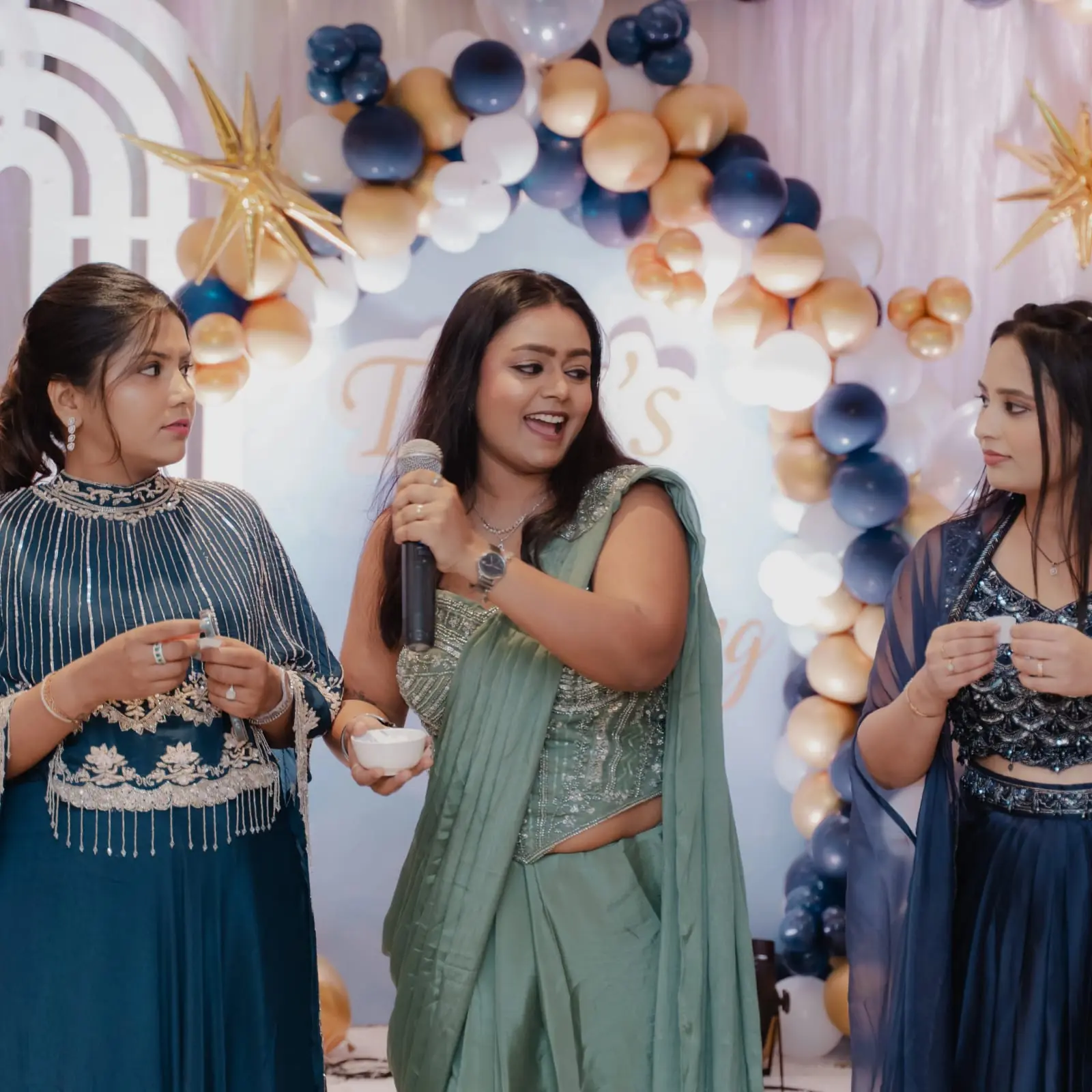 Palak Shah with a microphone, flanked by two guests at a celebration under a navy and gold balloon arch