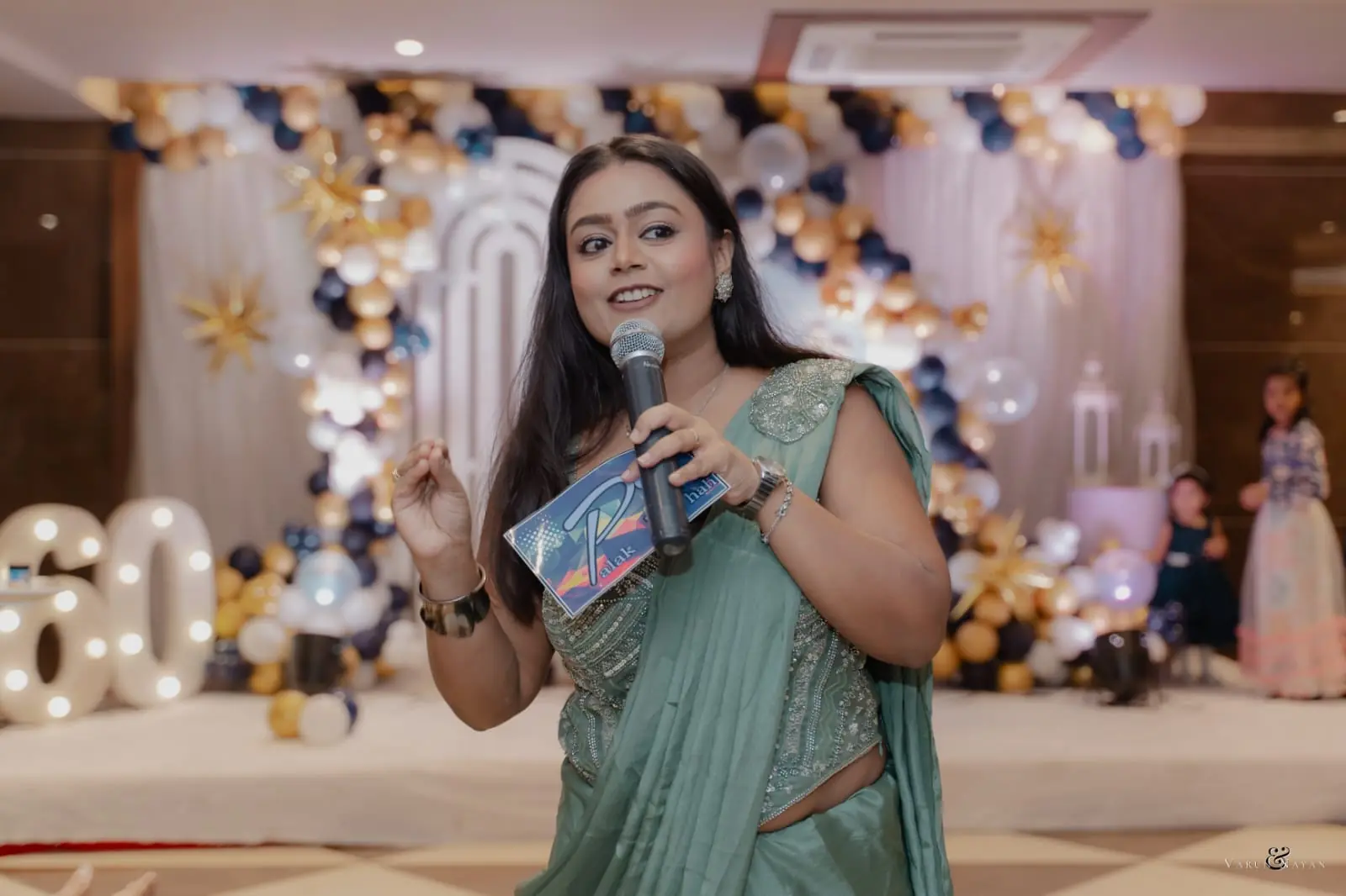 Palak Shah hosting a celebration, microphone in hand, before a navy and gold balloon backdrop