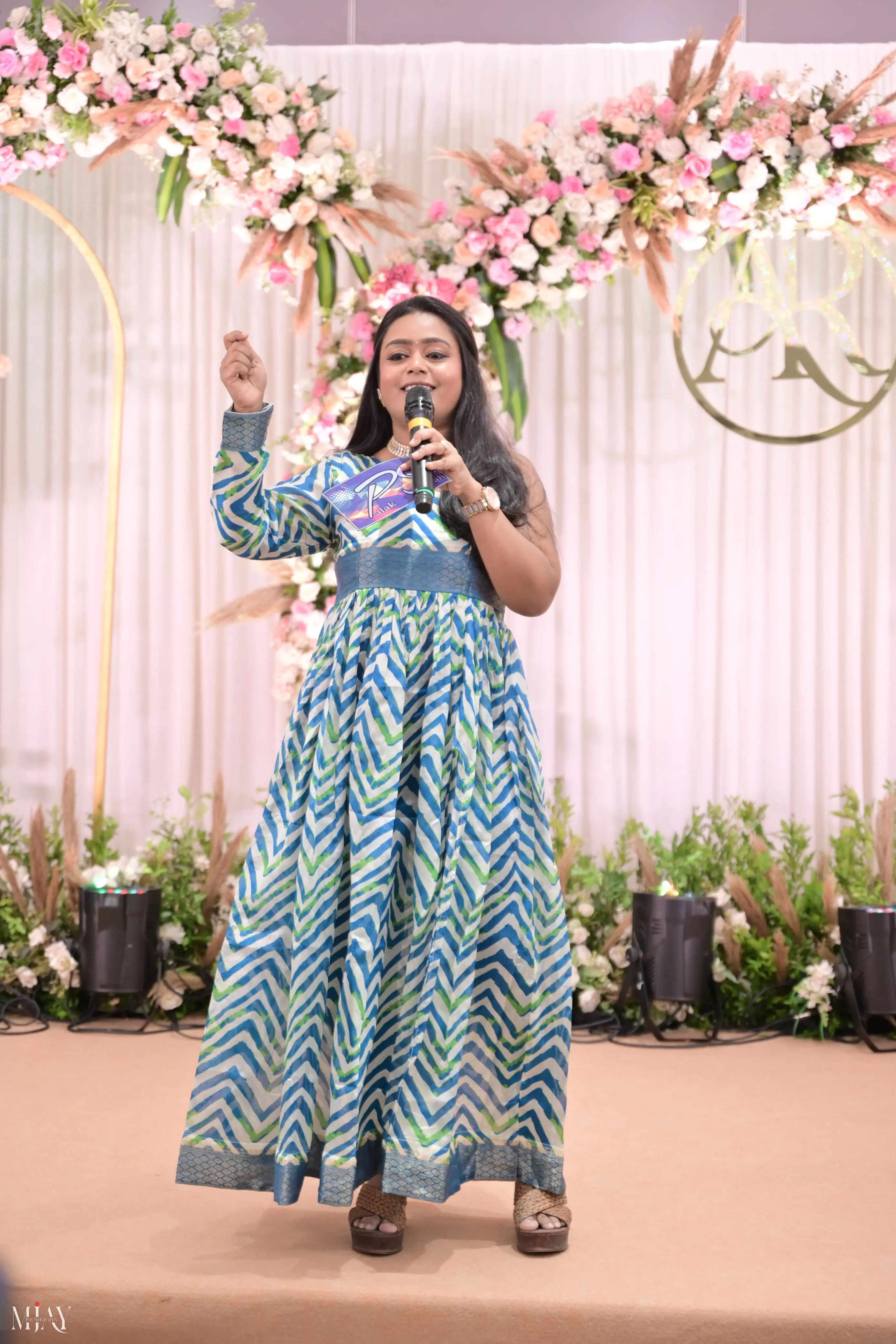 Palak Shah on a wedding stage, microphone in hand, in a blue and white chevron-print gown before a pink floral backdrop
