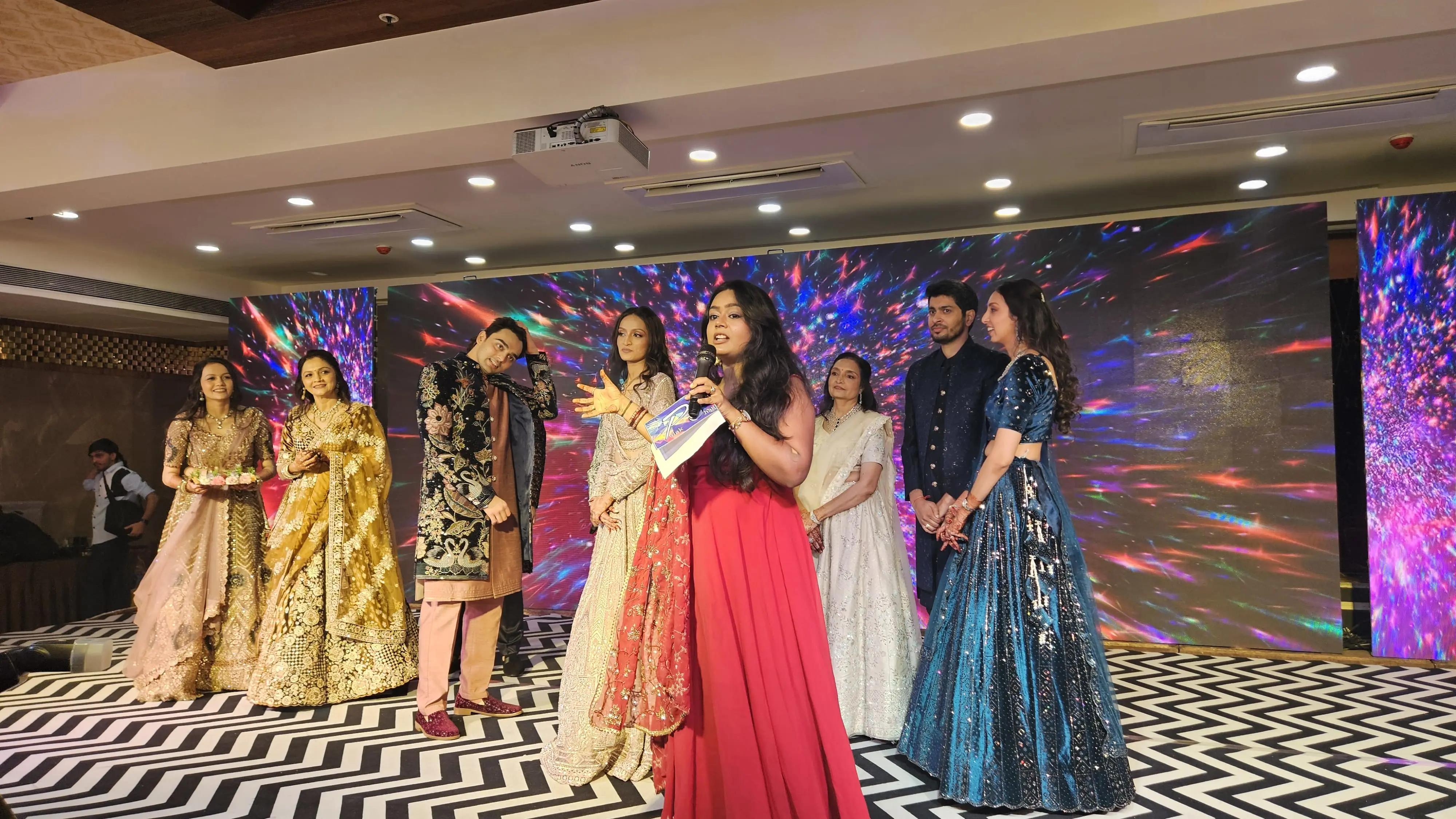 Palak Shah in a red gown, microphone in hand, addressing guests on a stage beside a family in festive attire, with a colourful light backdrop